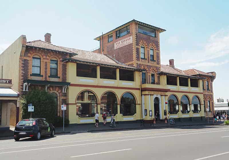 The historic Vue Grand hotel and restaurant in Queenscliff with people walking by and a parked car.