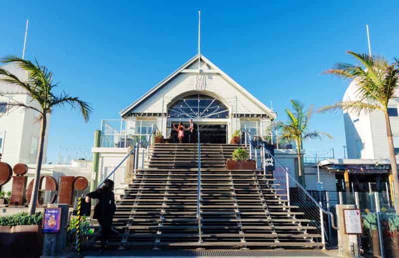 Stunning white building of the Geelong pier restaurant Wah Wah Gee with stairs, bright blue sky and green palm trees.